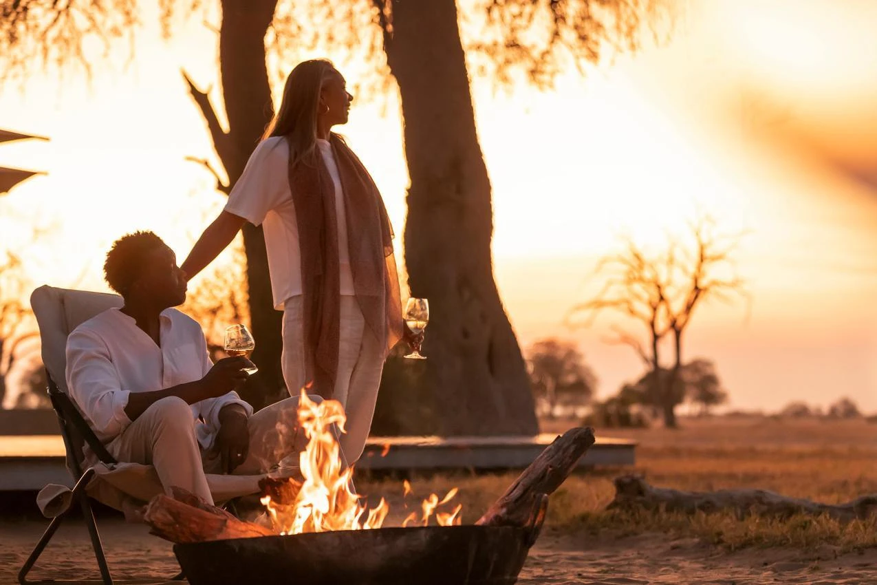 Couple enjoys an African photo safari with wine by a campfire at sunset.