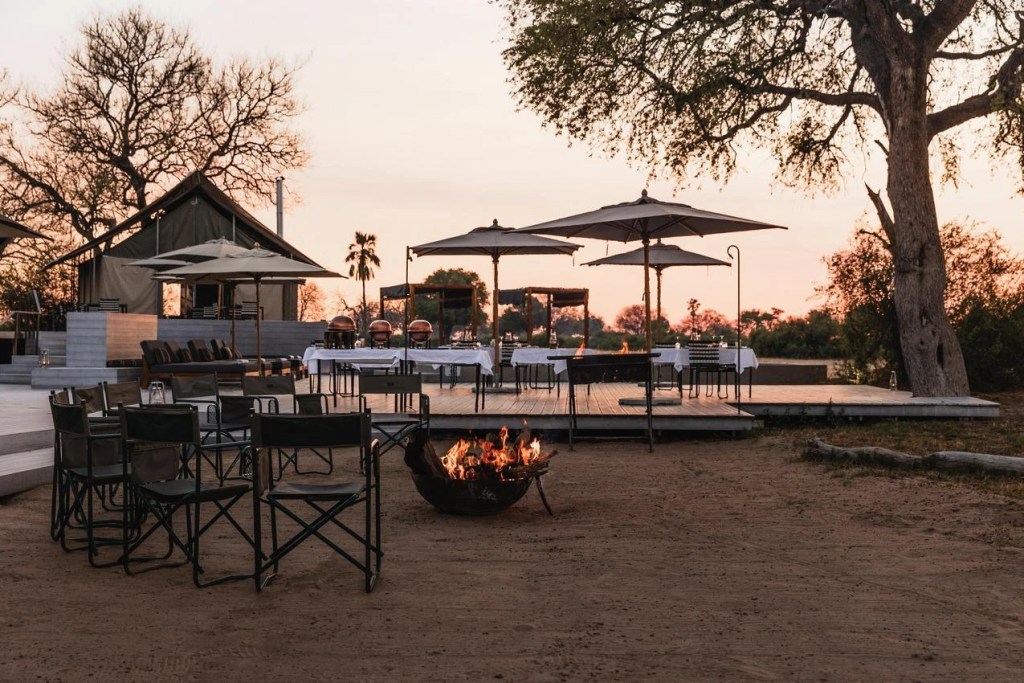 African safari camp with tables set for dinner, fire pit, and luxury tents at sunset. Photo safari experience.