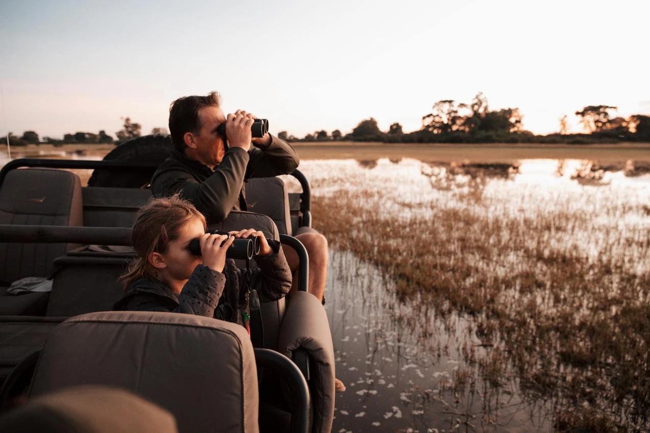 Father and daughter on African photo safari using binoculars, water reflection.