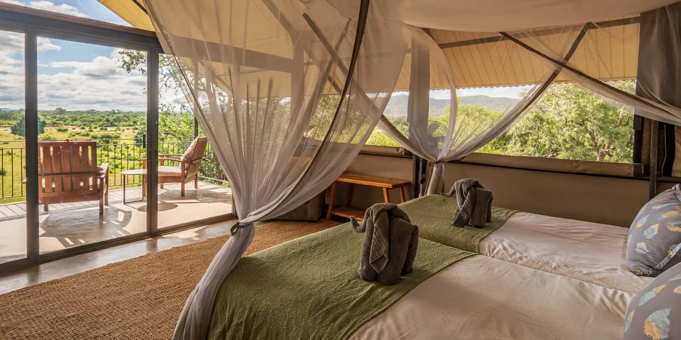 Safari tent interior with twin beds, elephant towel art, and a balcony overlooking the African savanna. Photo safari lodging.