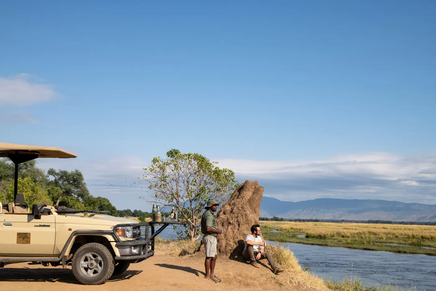 African photo safari: Safari jeep, guide, and traveler by river, mountains in background.