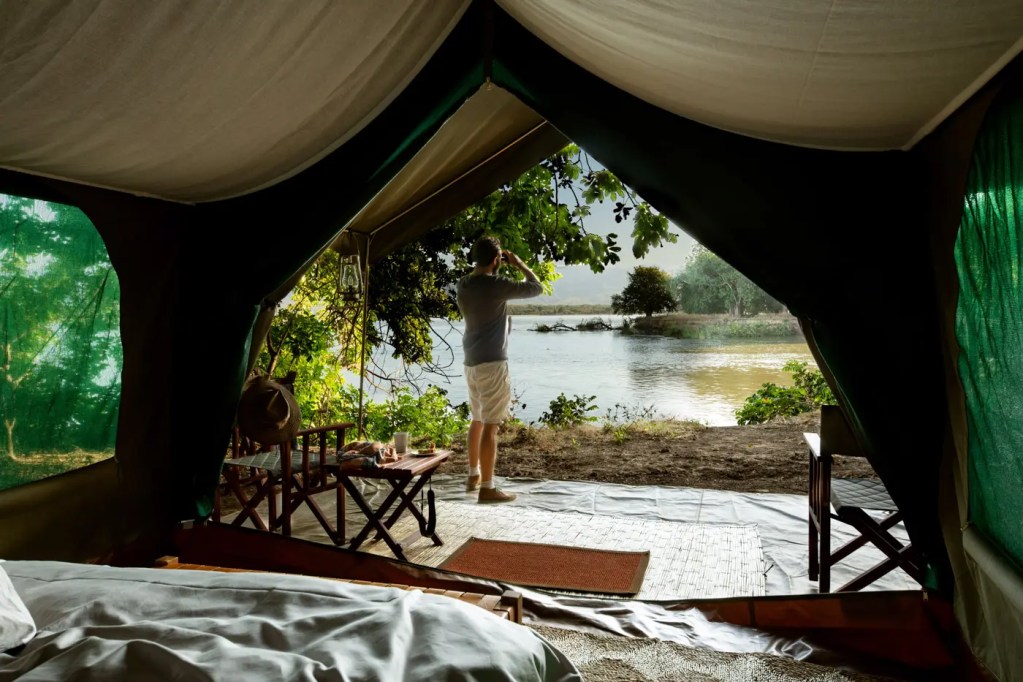 African photo safari tent view: Man enjoys river view. Luxury safari camping.