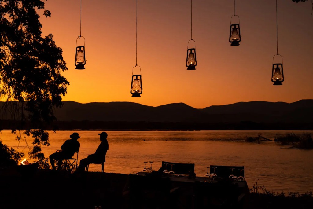 African photo safari: Silhouetted couple by lake at sunset, lanterns above. African safari landscape.
