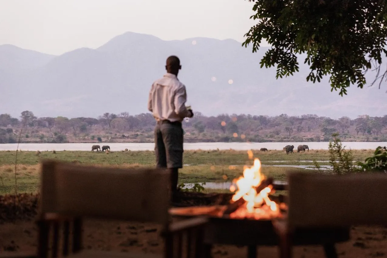 African photo safari: Man watches elephants from a campfire near a river with mountains.