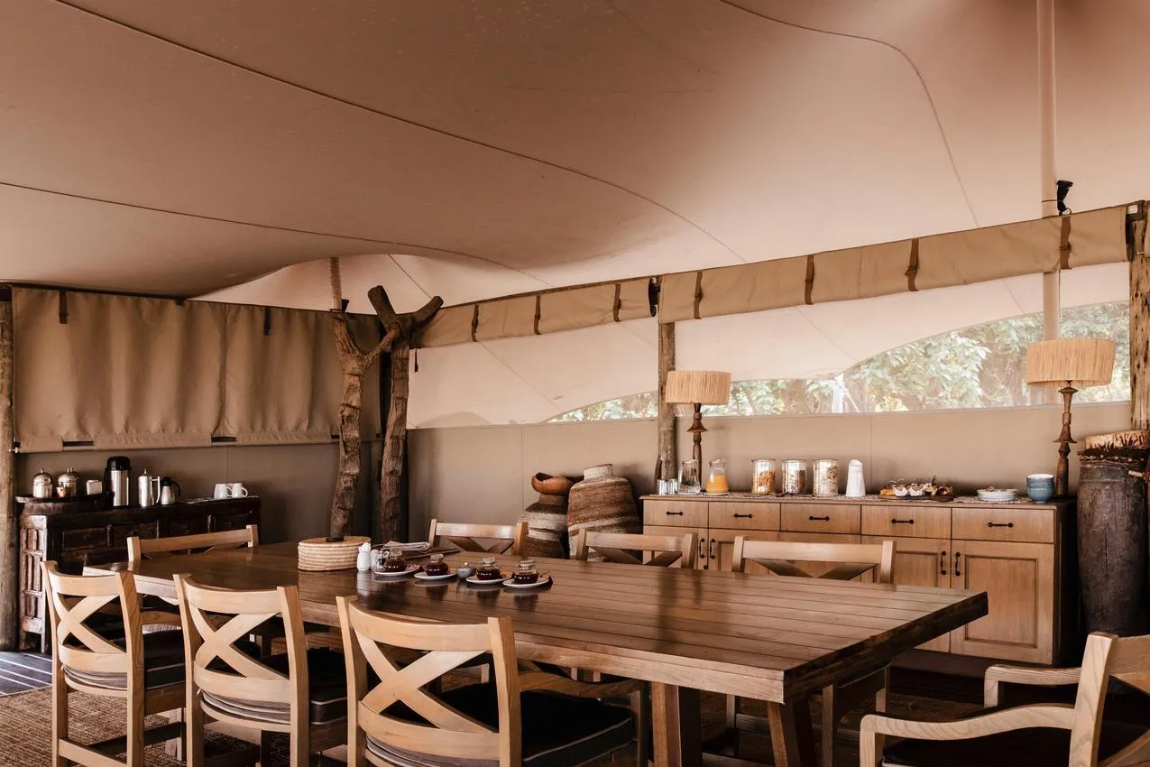 African safari dining tent with a long wooden table and chairs, buffet, and natural light. Photo safari experience.