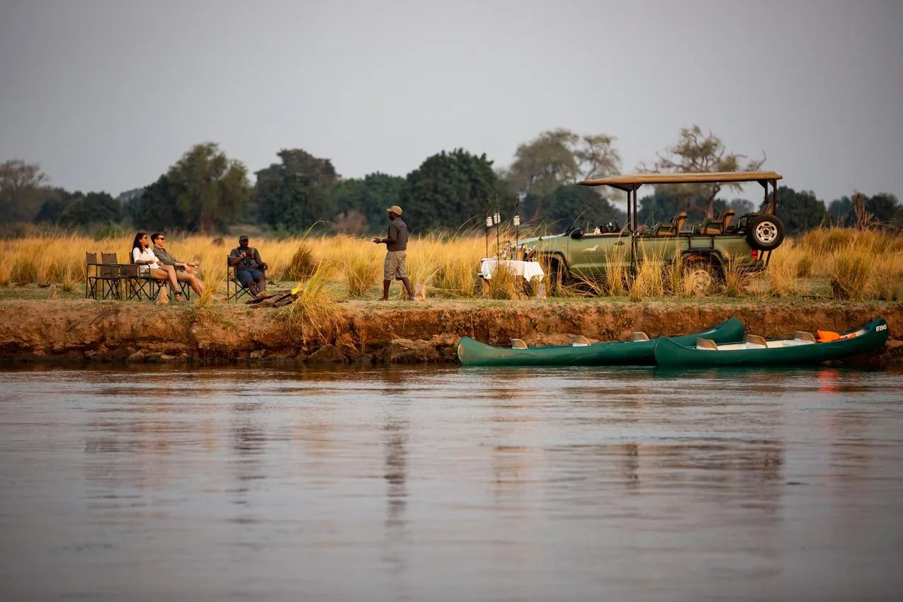 African photo safari: People relaxing by the river with safari vehicle and canoes.