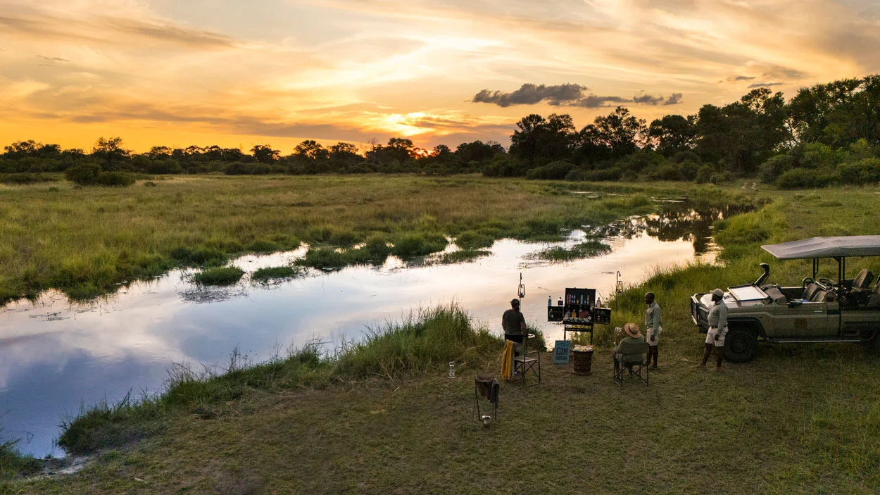African photo safari at sunset: People gather by a river with a safari vehicle, enjoying the scenic landscape.