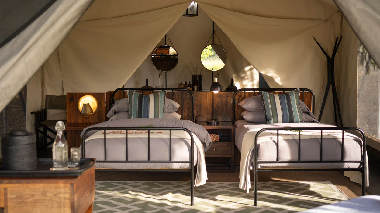 Safari tent interior with two beds, wood headboards, and African-inspired decor. Luxury photo safari accommodation.