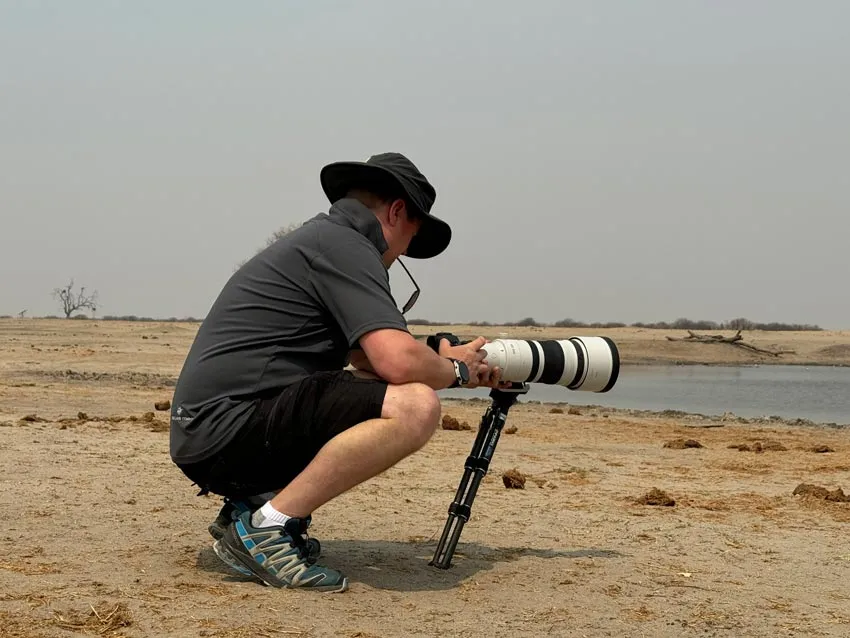 African photo safari: Photographer with telephoto lens on tripod, crouching near watering hole. About photo safari company.