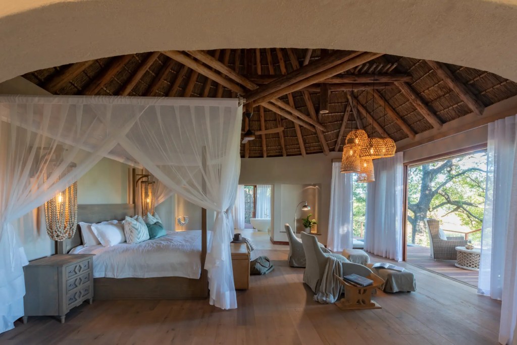 Luxury African safari lodge bedroom with canopy bed, thatched roof, and scenic views.