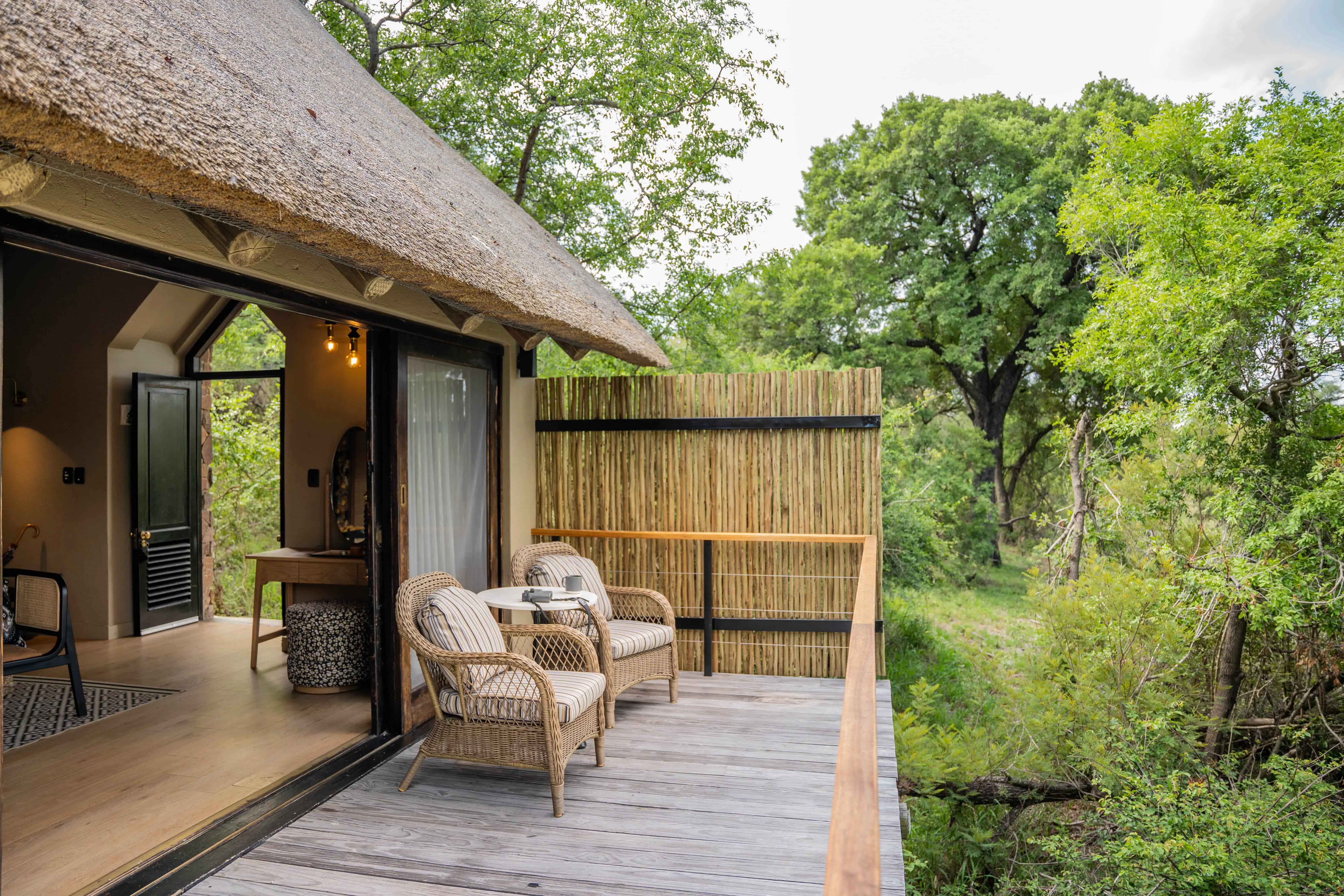 African safari lodge balcony with thatched roof, chairs, and lush greenery.