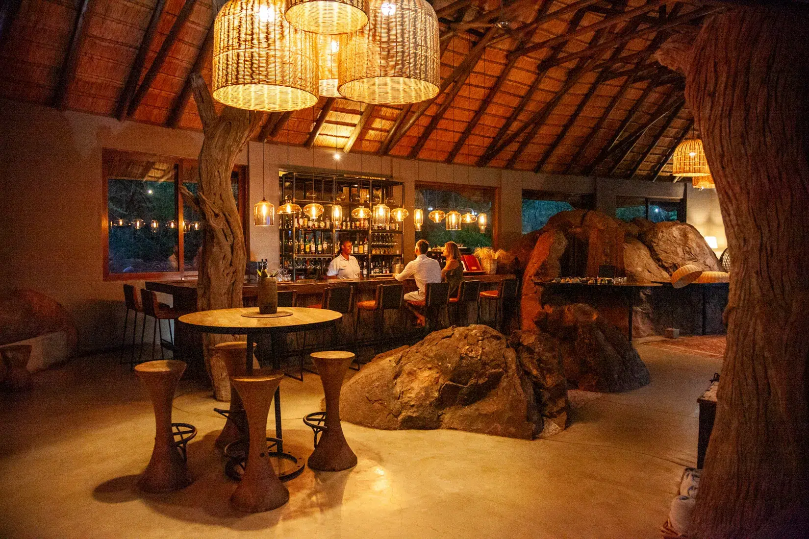 Interior of a luxury African safari lodge bar with thatched roof, guests at bar.