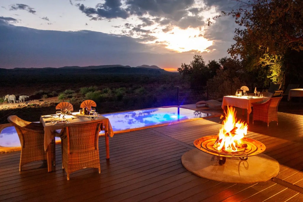 Luxury African safari lodge dining area with pool, fire pit, and elephants at sunset.