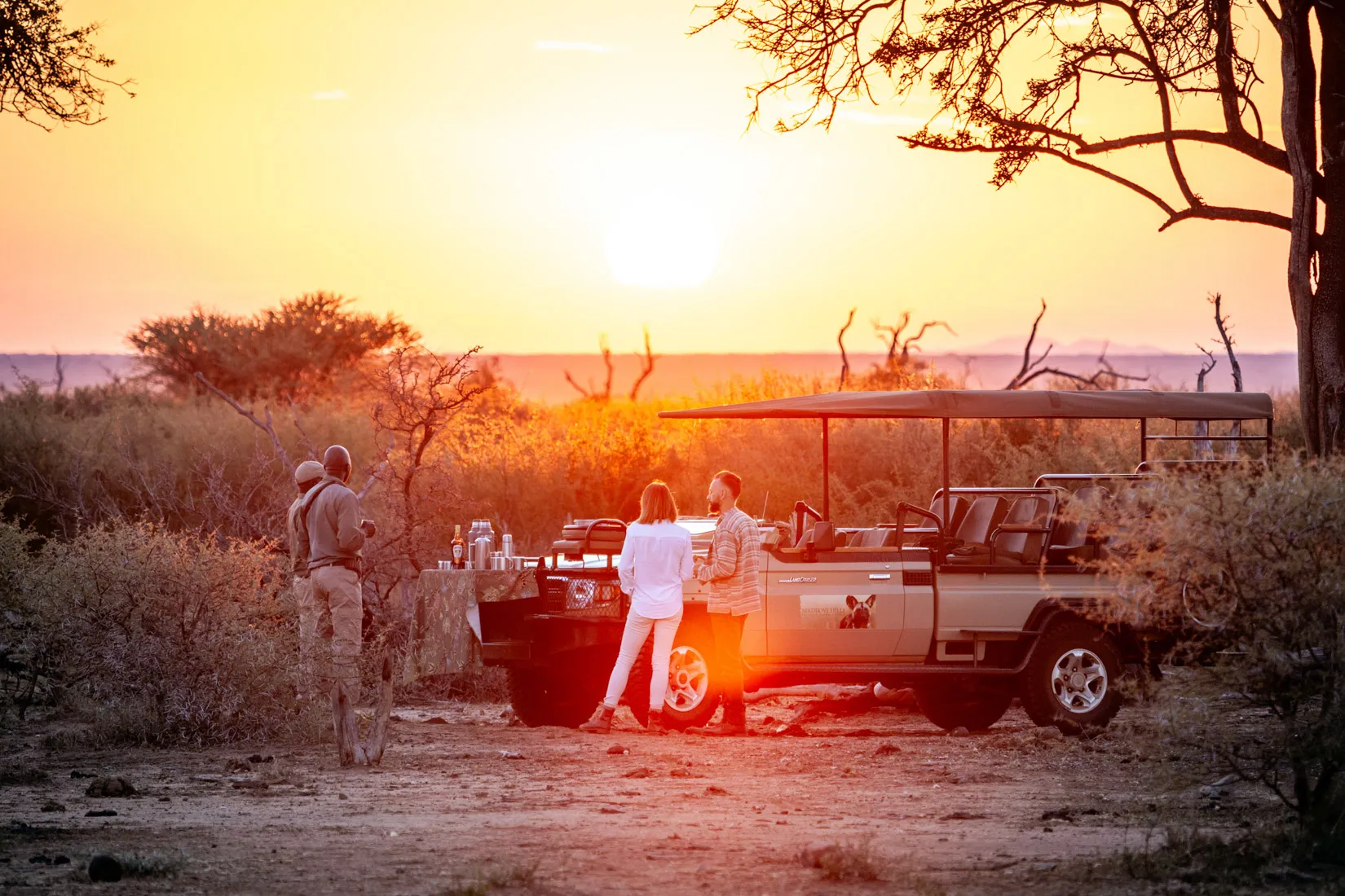 African safari sunset with guests enjoying drinks near a safari vehicle in Madikwe